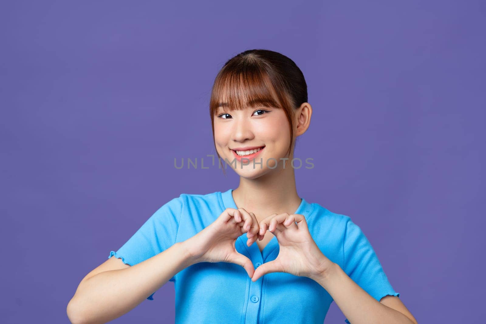 Young asian woman shows her fingers heart-shaped sign and presses to chest on purple background.