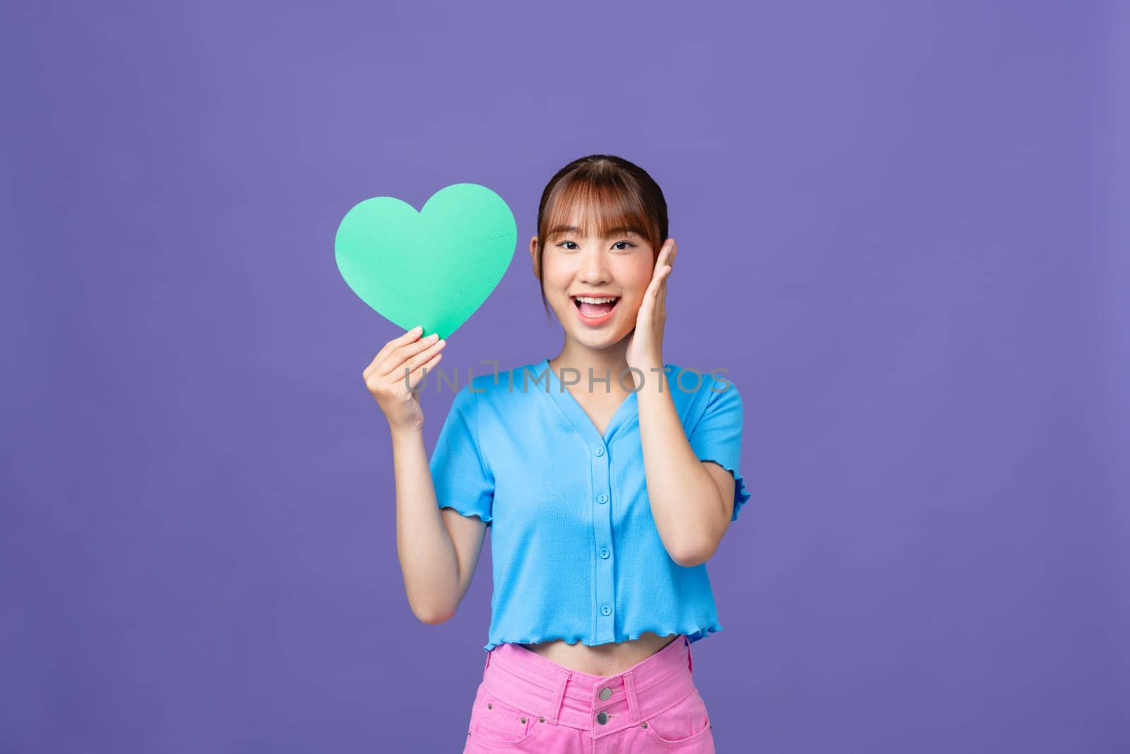 Cheerful young woman hold hands green heart paper shape isolated on purple background by makidotvn