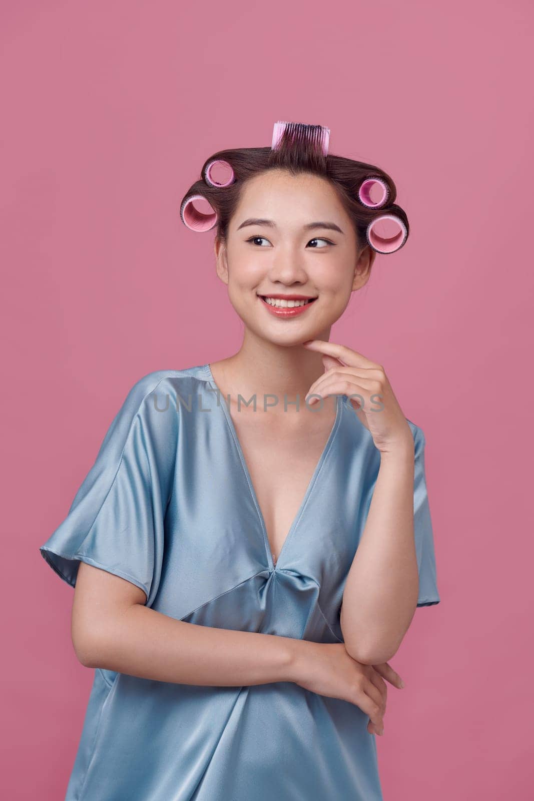 Happy young woman in blue dress and hair curlers on pink background by makidotvn