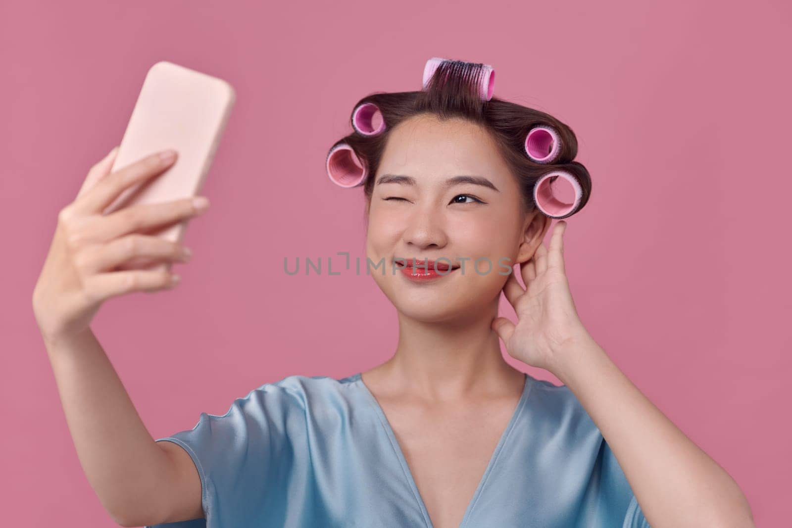 cheerful woman applies hair curlers posing in smartphone camera dressed over pink background.