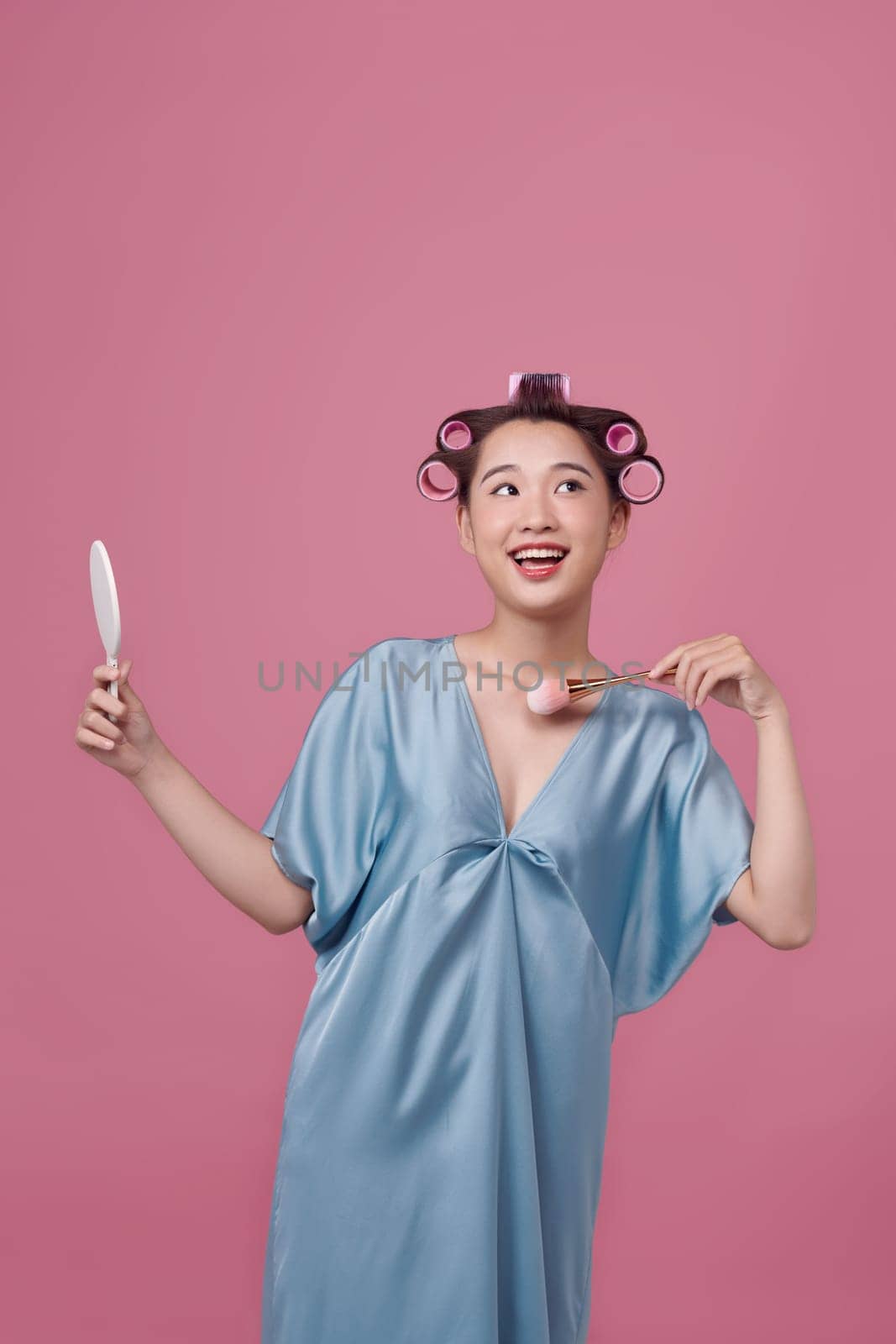 Cute girl with hair curlers doing makeup on her face at the makeup table, using makeup brush