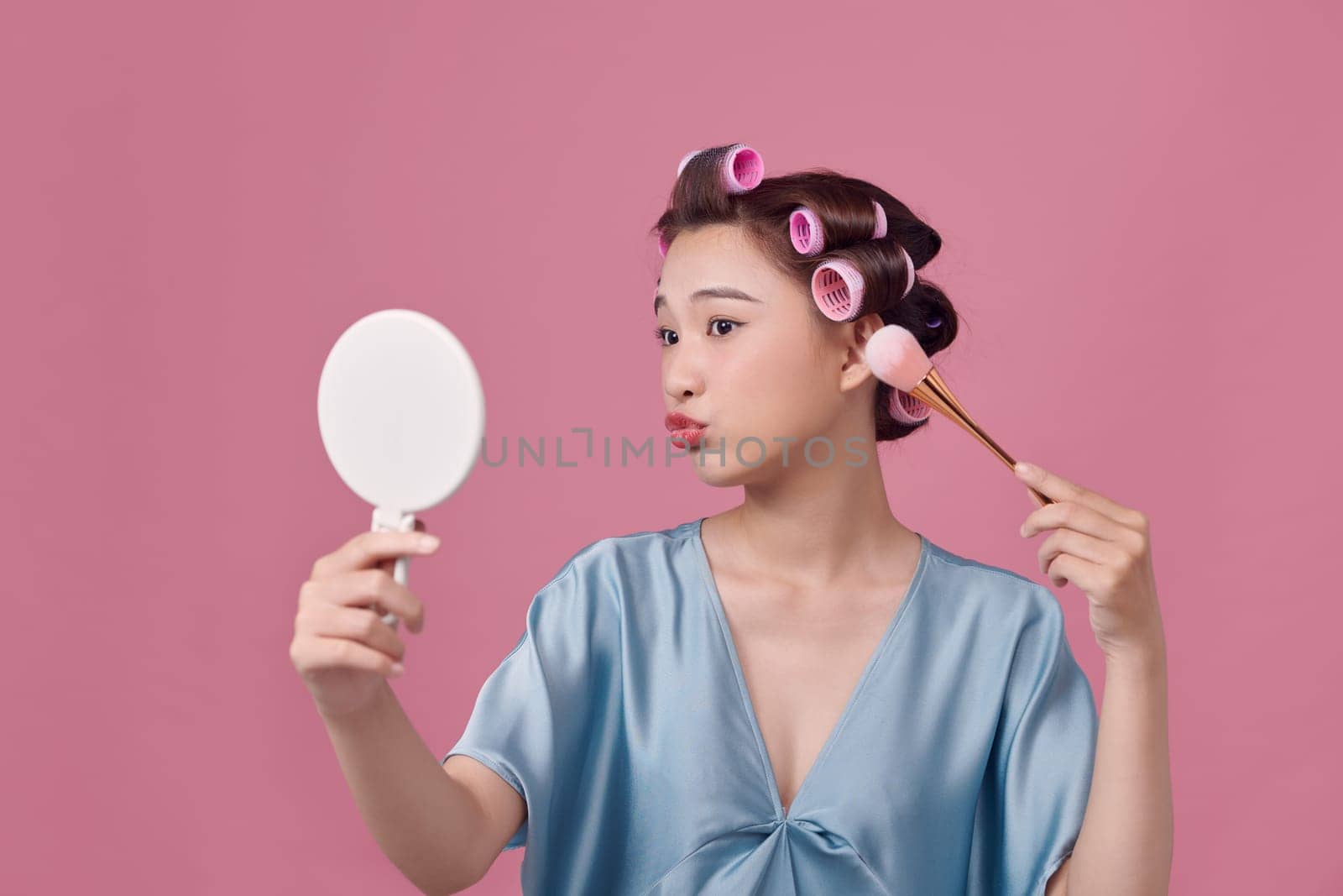 Pretty woman with curlers on hair, holding makeup brushes and mirror in hands  by makidotvn