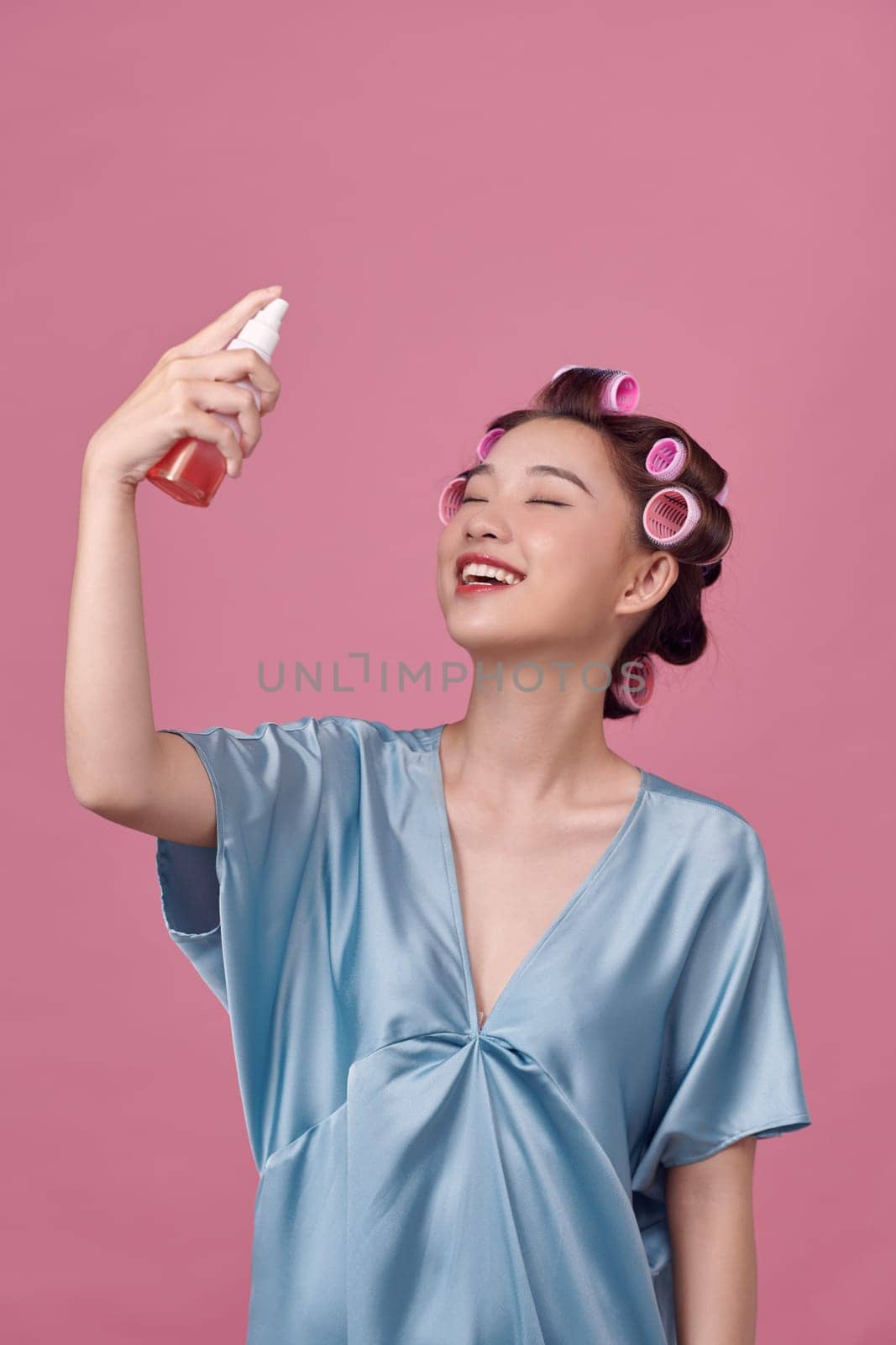Beautiful Asian woman applying spray water on face  on pink background with curlers in her hair by makidotvn