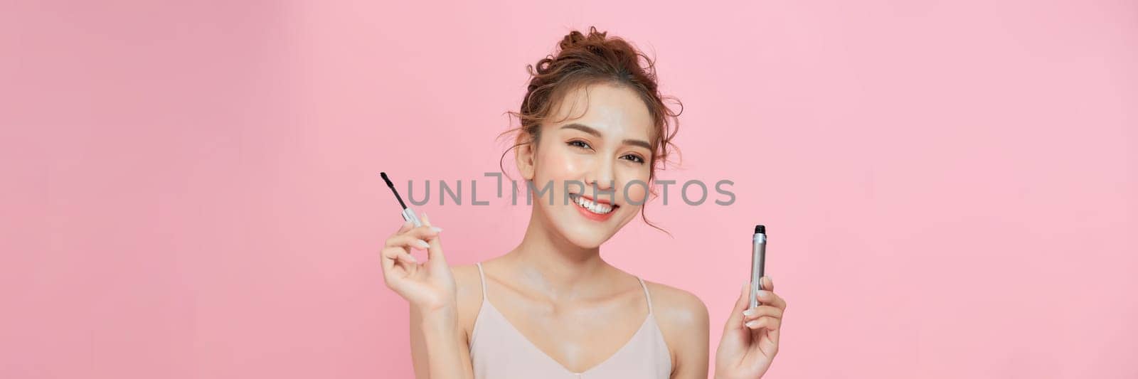 happy young woman holding mascara tube and brush isolated on pink background by makidotvn