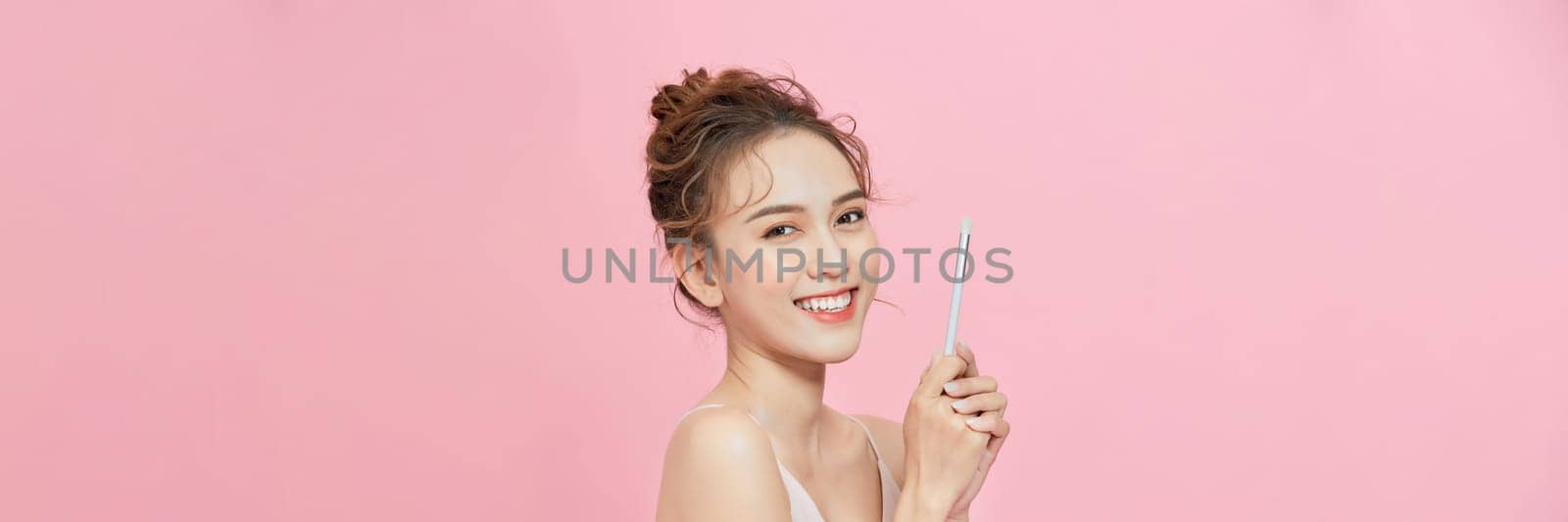 Smiling young asian woman holding blush brush near face isolated on pink