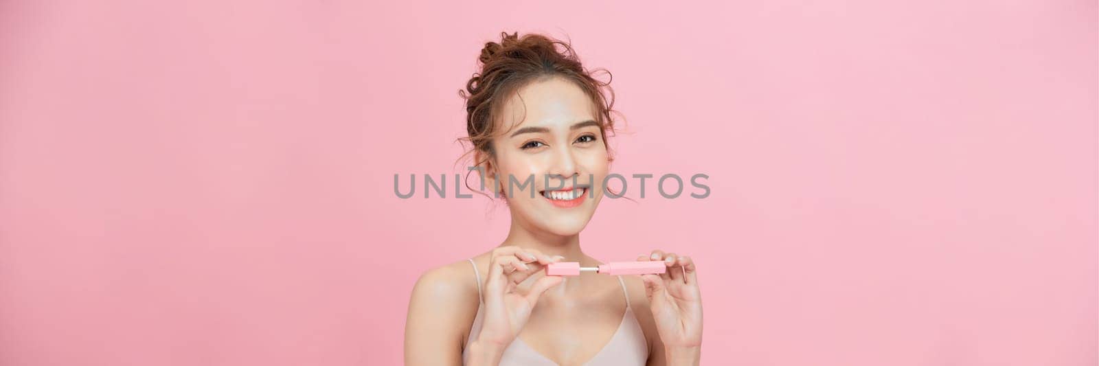 Young woman using lip gloss looking at camera on pink studio background by makidotvn