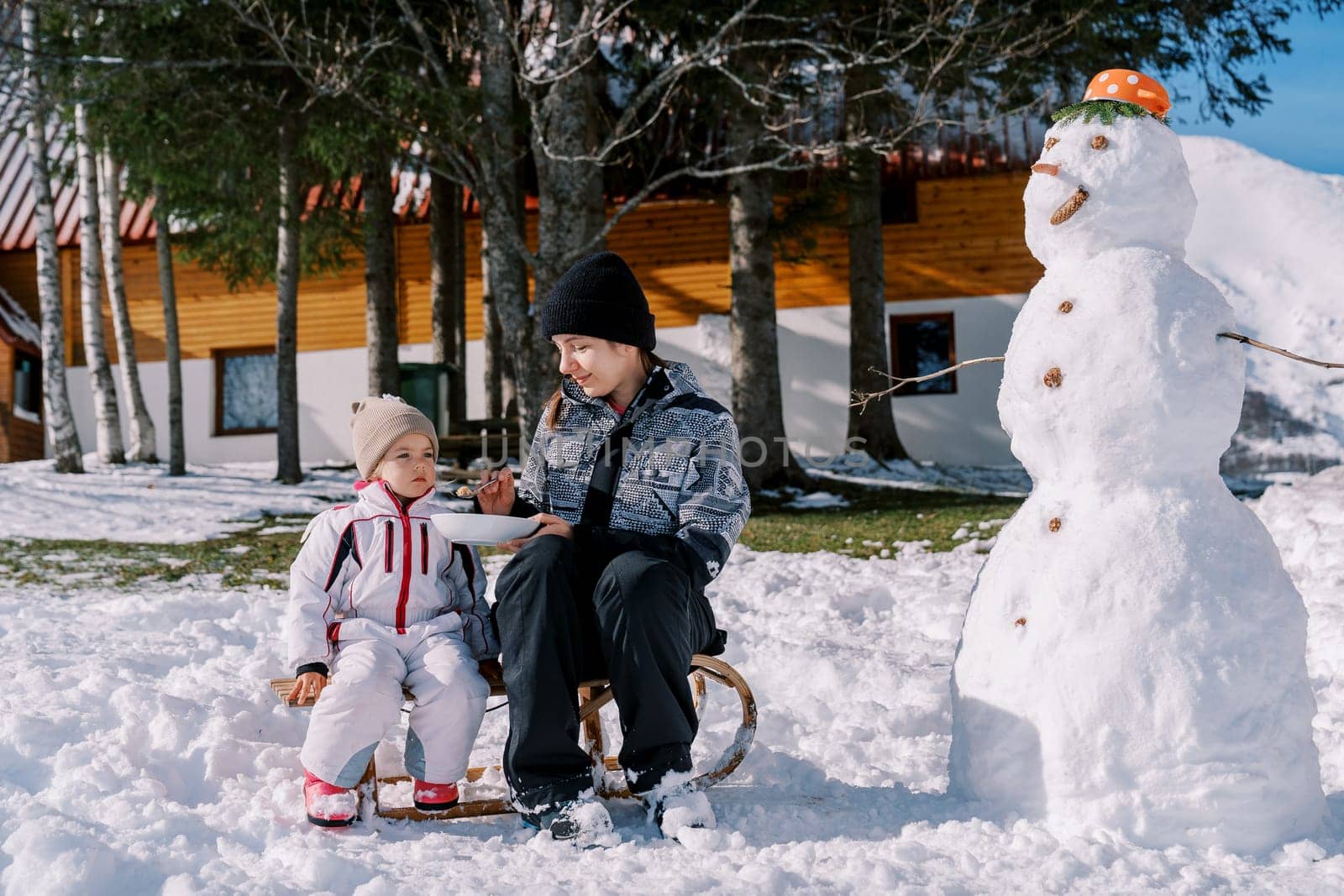 Mom feeds a little girl porridge from a spoon, sitting on a sleigh near a snowman in the yard by Nadtochiy
