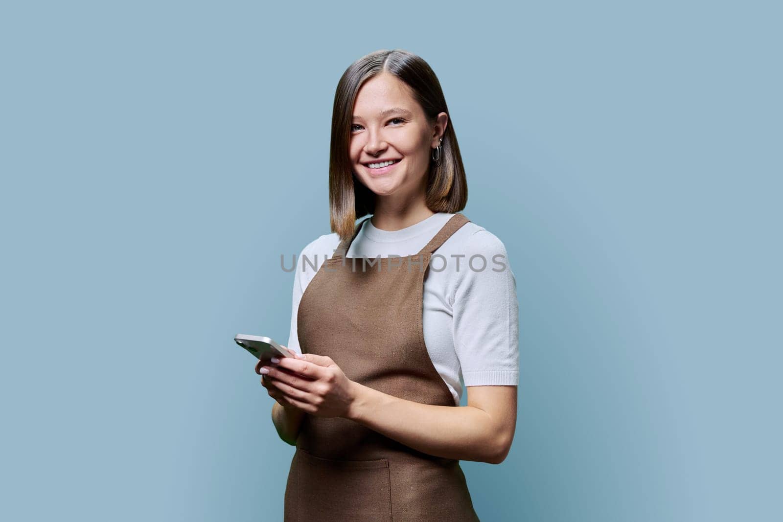 Portrait of young woman in apron holding smartphone in hands on blue background by VH-studio