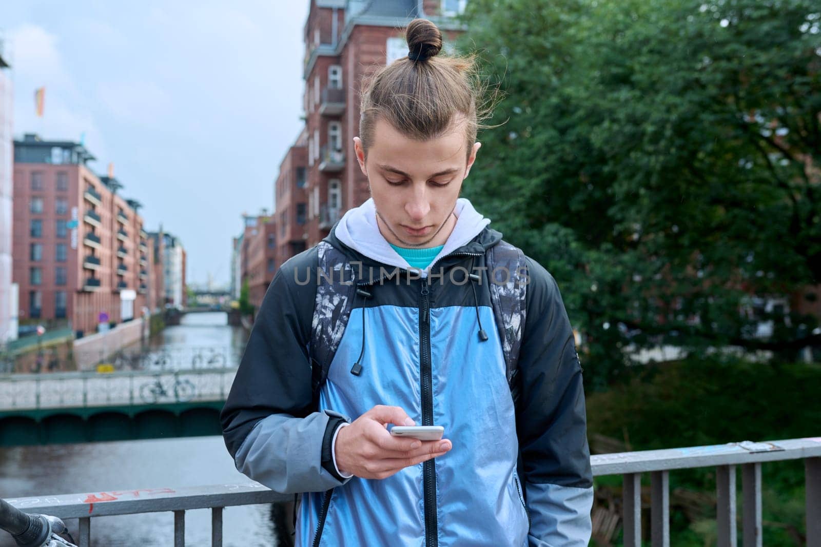 Fashionable young male with backpack using smartphone outdoors in city. Hipster with trendy hairstyle beard with phone in hands mobile applications for tourism travel communication entertainment study