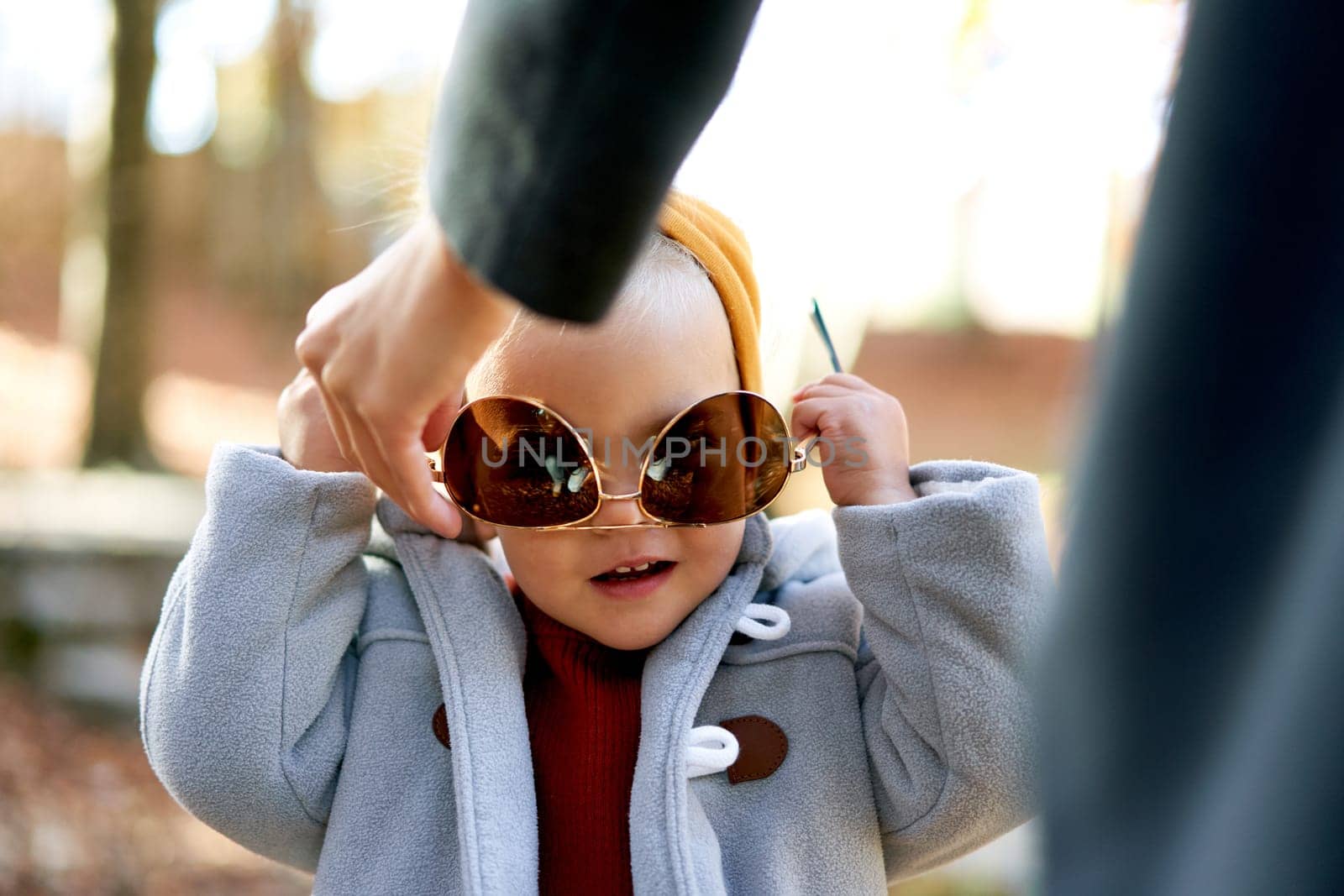 Little girl tries on her mother sunglasses while standing with her in the park. Cropped by Nadtochiy