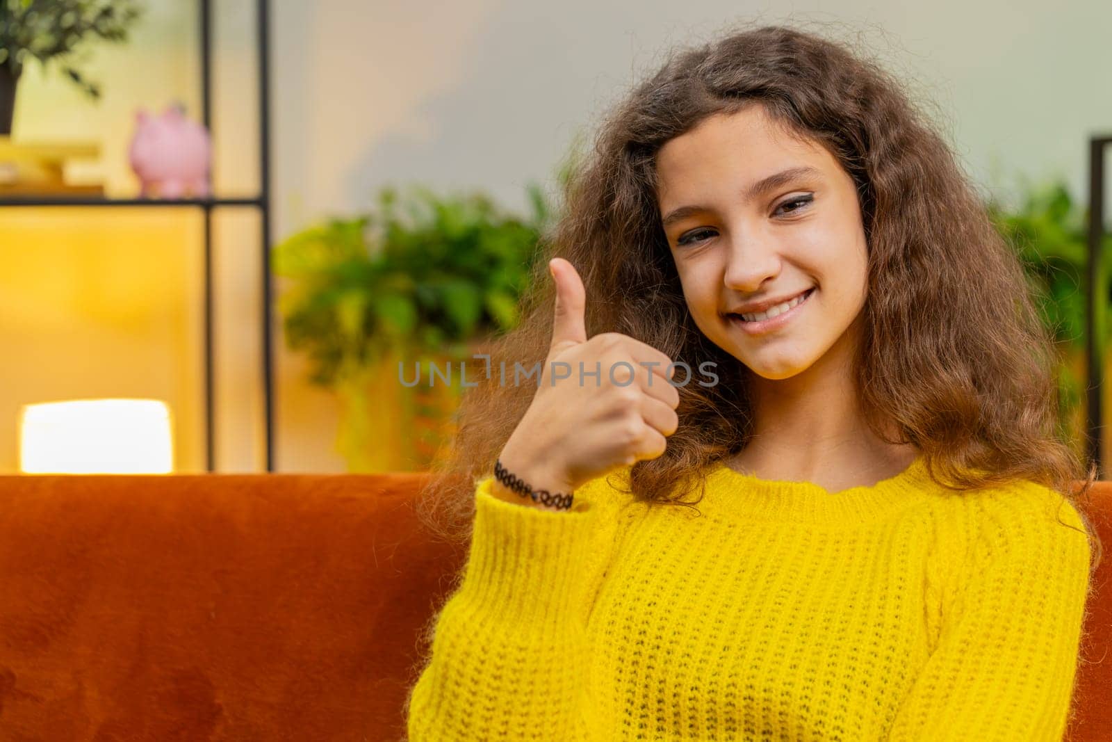 Like. Happy young Caucasian child girl looking approvingly at camera showing thumbs up like sign positive something good, positive feedback. Female teenager kid at home in room sots on sofa. Vertical