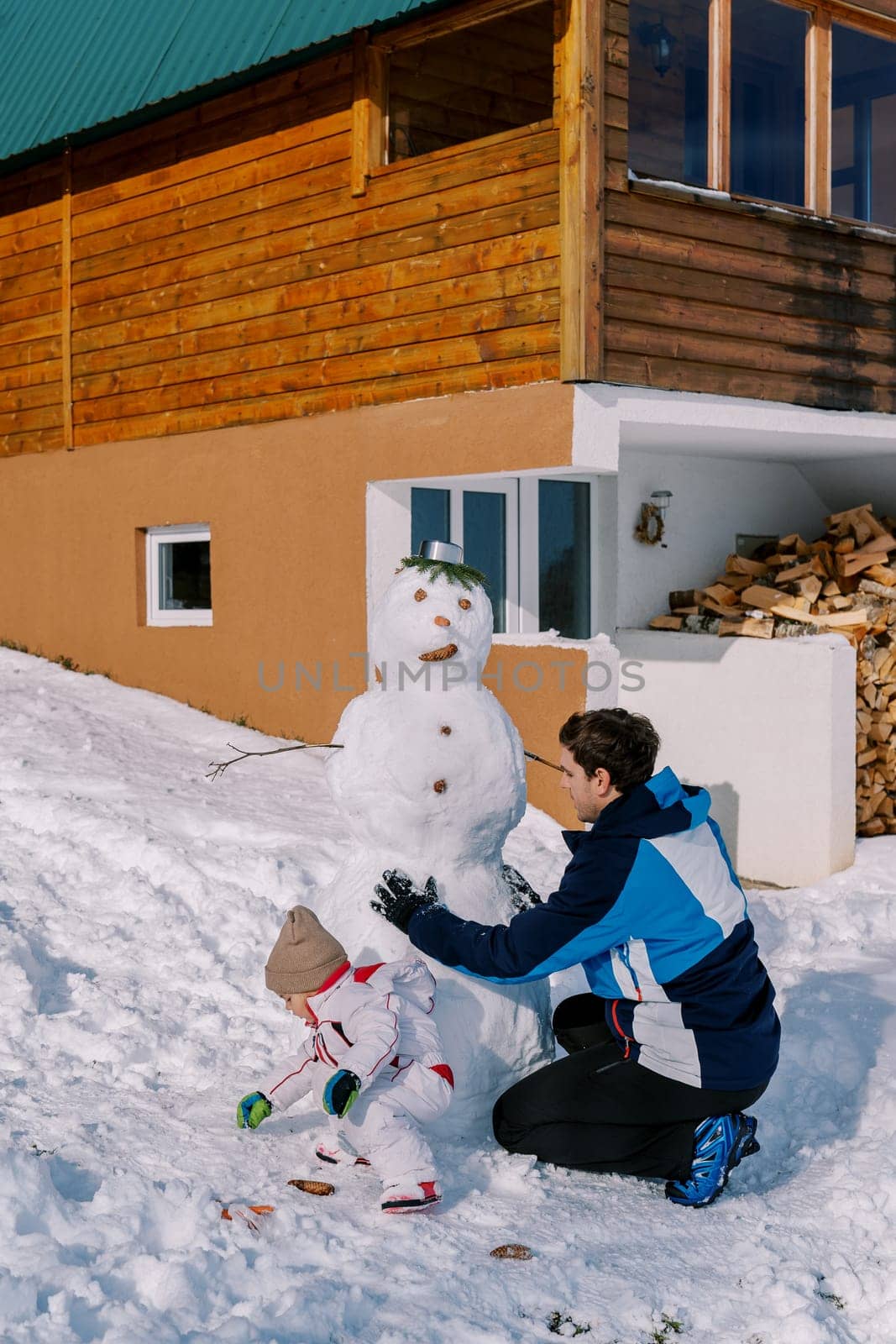 Dad and little girl decorate a snowman with buttons near a woodpile of a wooden chalet in the snow. Back view by Nadtochiy