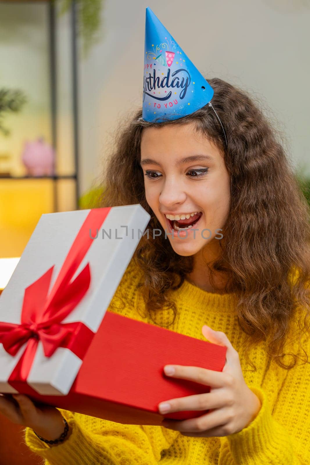 Happy 14-15 years child girl wears festive birthday cap hat hold gift box with ribbon congratulating. Teenager kid celebrating party event opening delivery greeting present at home room. Vertical