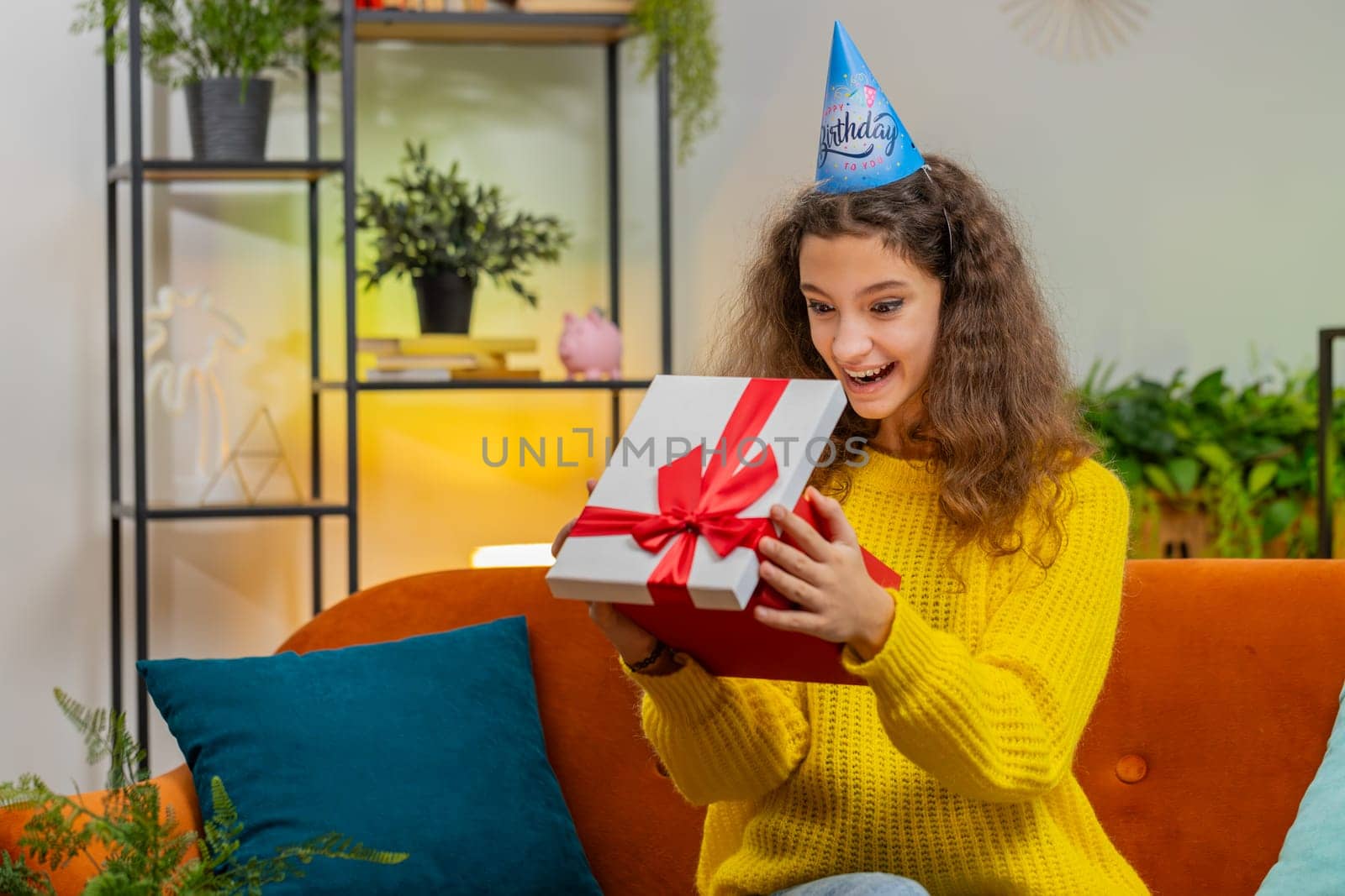 Happy 14-15 years child girl wears festive birthday cap hat hold gift box with ribbon congratulating. Teenager female kid celebrating party event opening delivery greeting present at home room on sofa