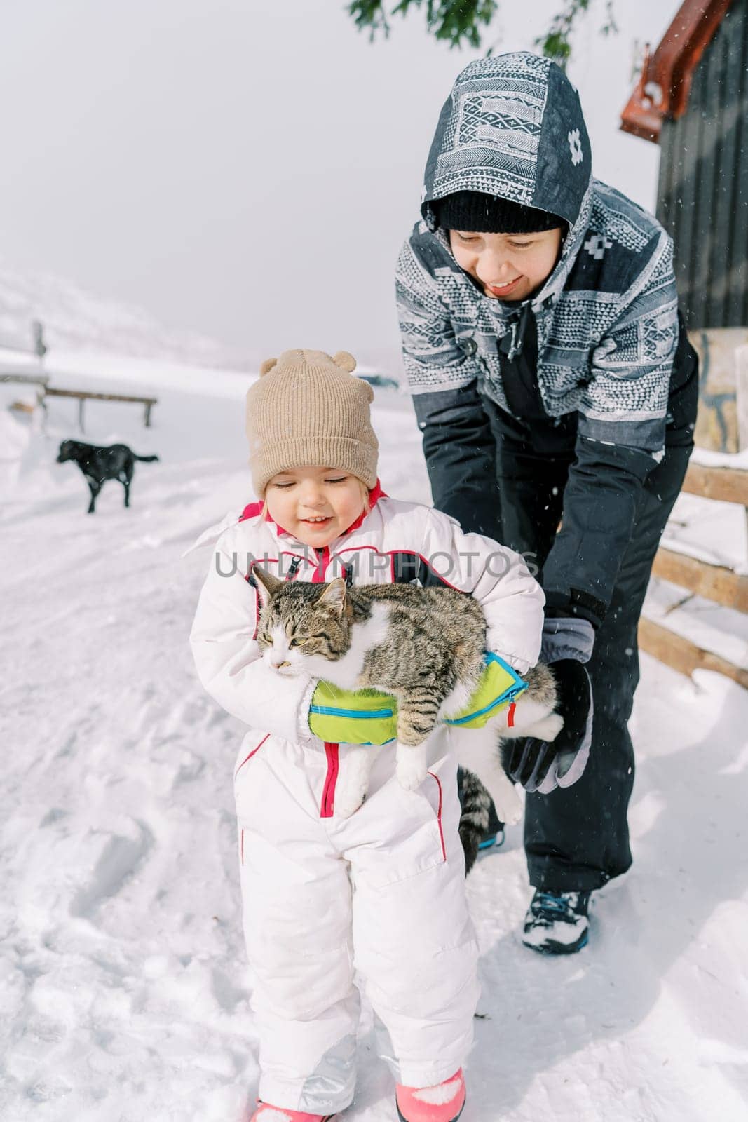 Smiling mother helps a little girl carry a tabby cat in her arms along a snowy road. High quality photo
