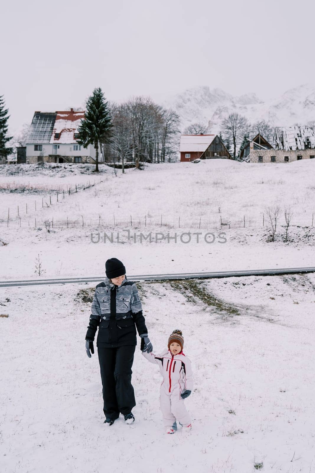 Mom and little girl walk along a snowy hill in the village, holding hands by Nadtochiy
