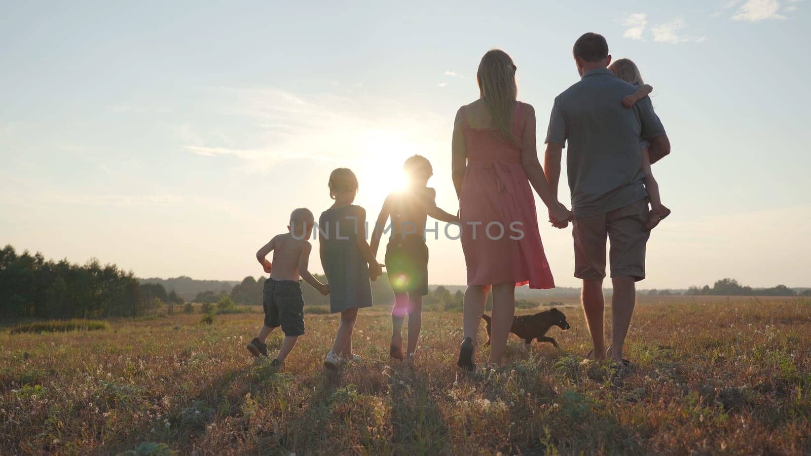 A friendly large family walks across the field at sunset with dog