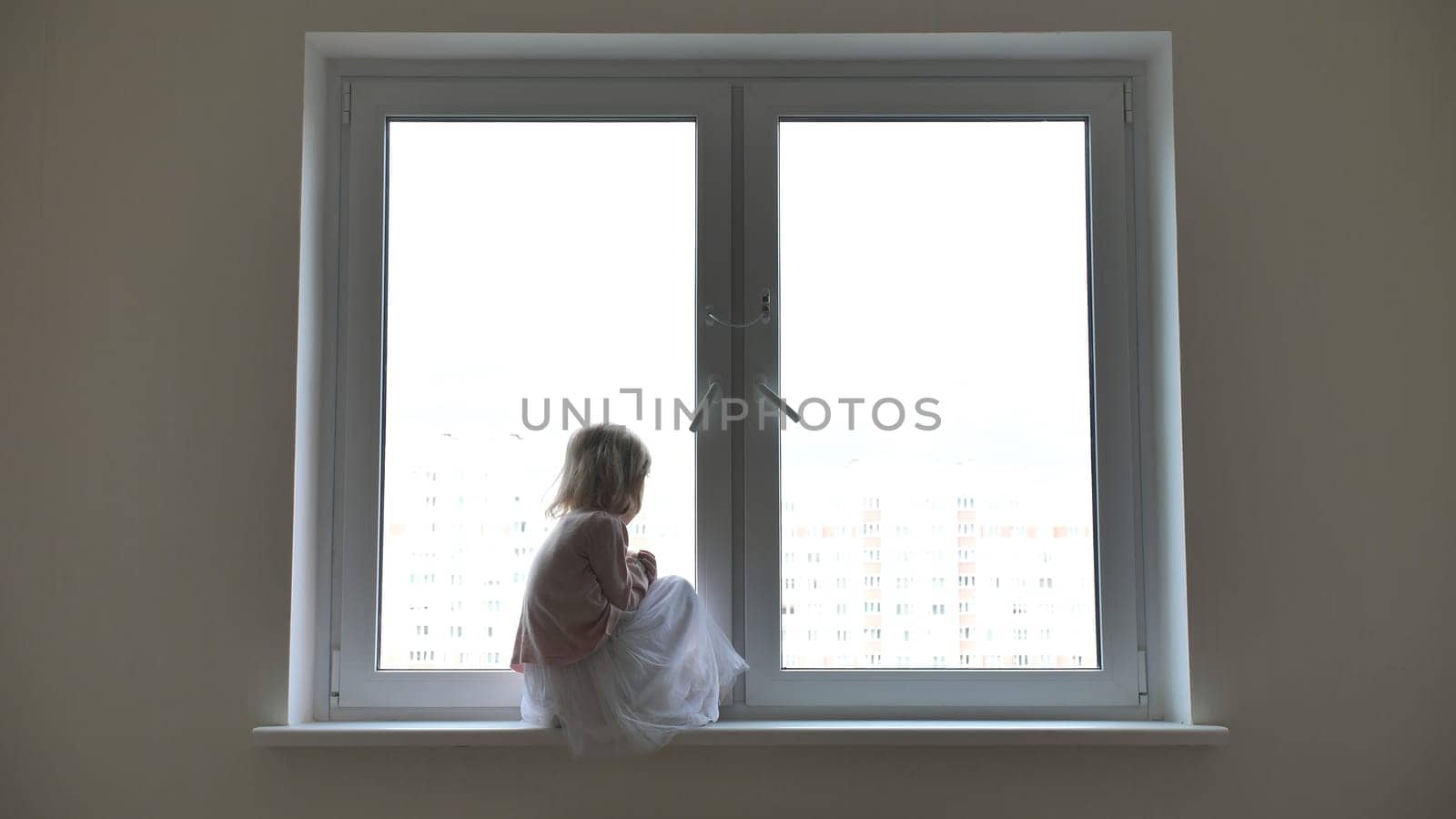 Lonely child girl sitting on the windowsill at the window in the apartmen