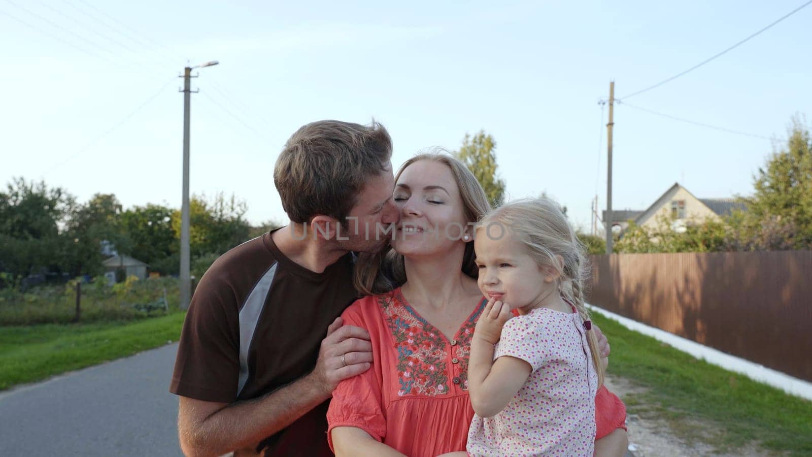 Young spouses with their daughter are walking around the village. The husband kisses his wife