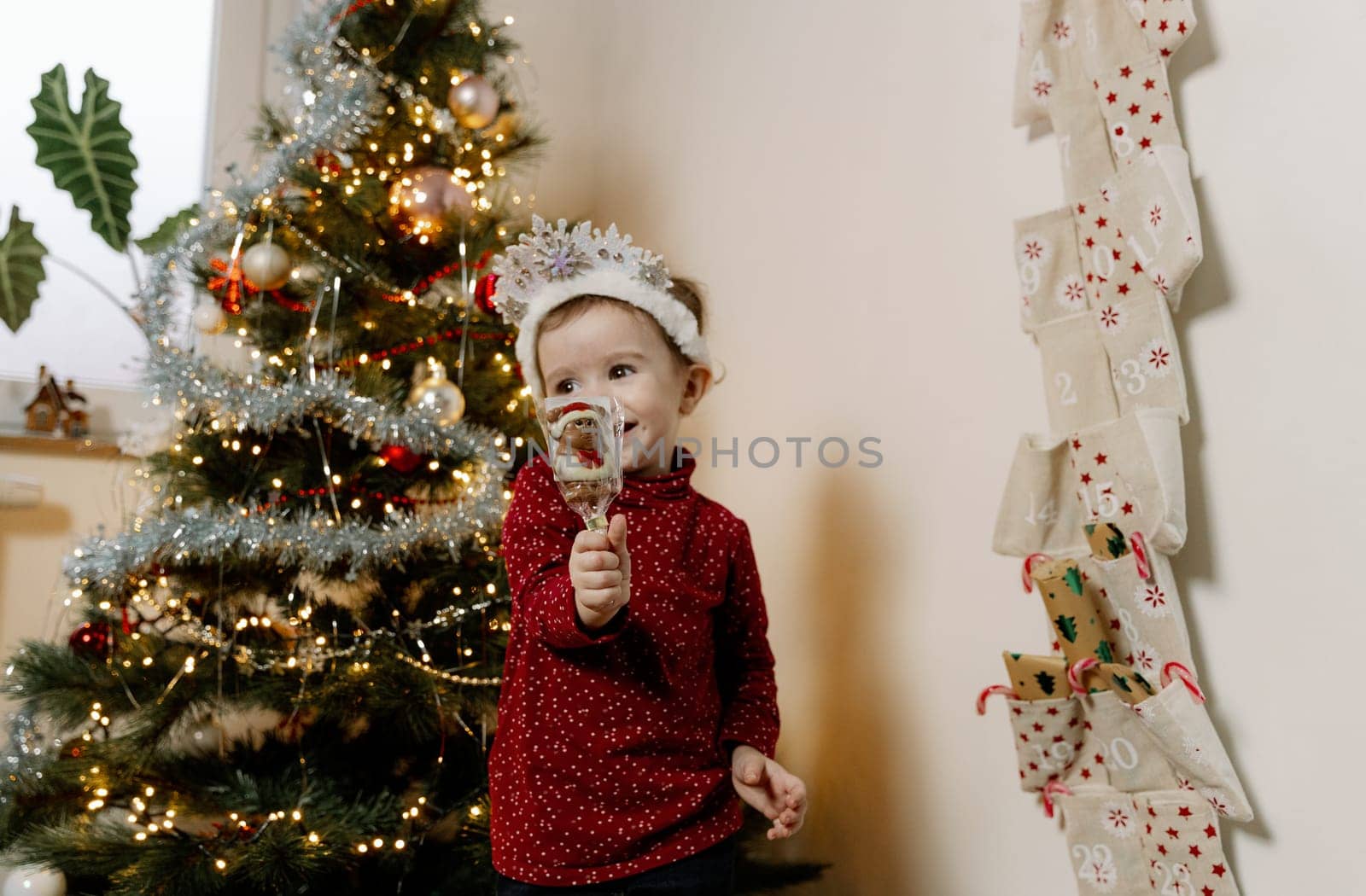 One little Caucasian girl with a white tiara of snowflakes stands between a Christmas tree and an advent calendar, holding a chocolate santa claus with one hand and covering one eye with it while standing in the living room, looking cutely to the side, close-up side view.