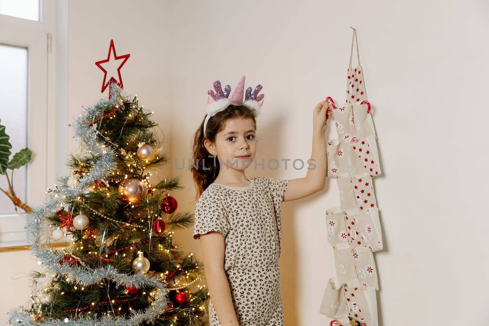 One beautiful Caucasian brunette girl with a unicorn horn headband stands by the Christmas tree and with her hand takes out a lollipop from a pocket with the number two of an advent calendar in the morning in the living room, close-up side view.