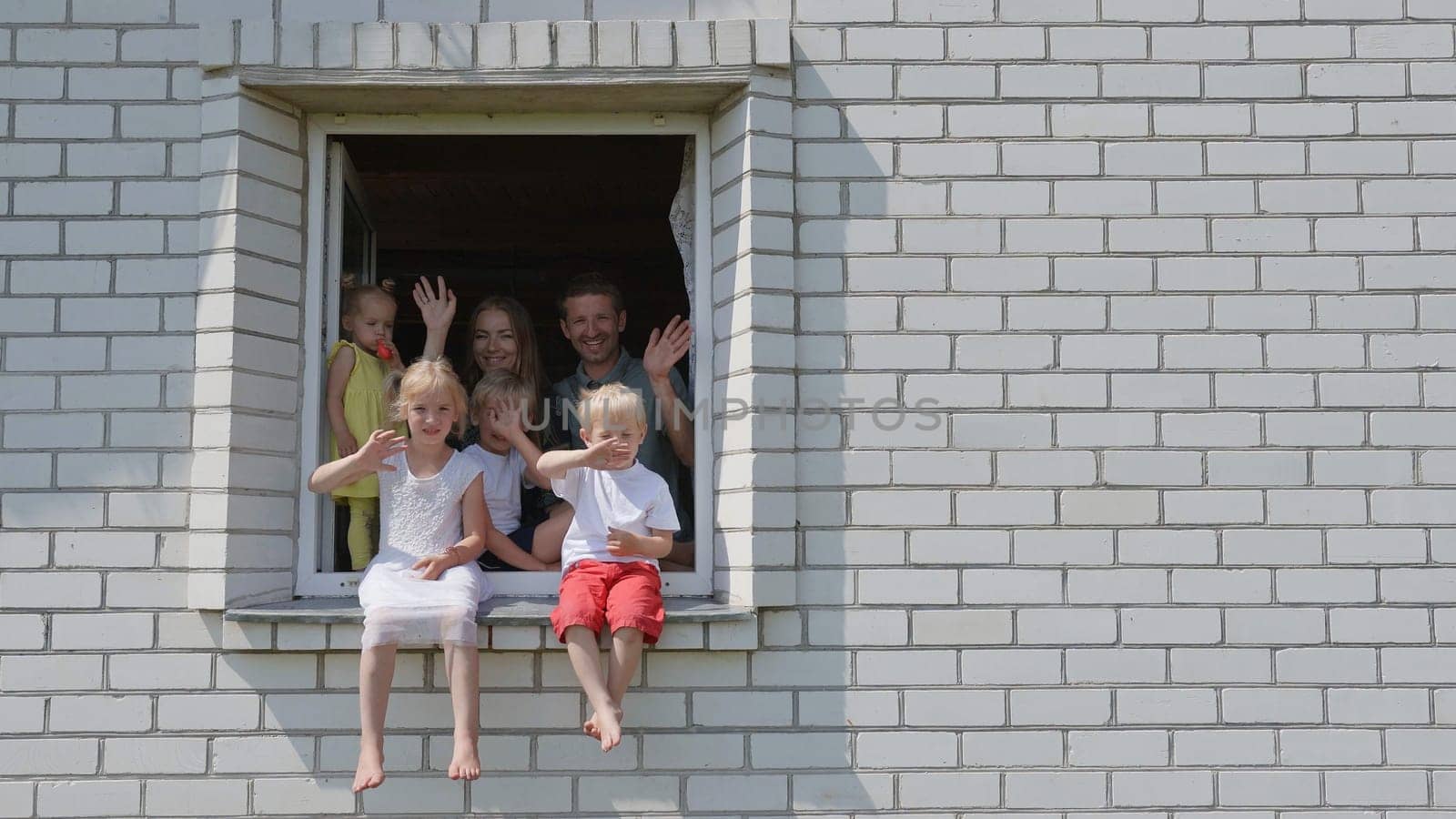 The concept of a large family. A family of 6 waves their hands from the window of their house. by DovidPro