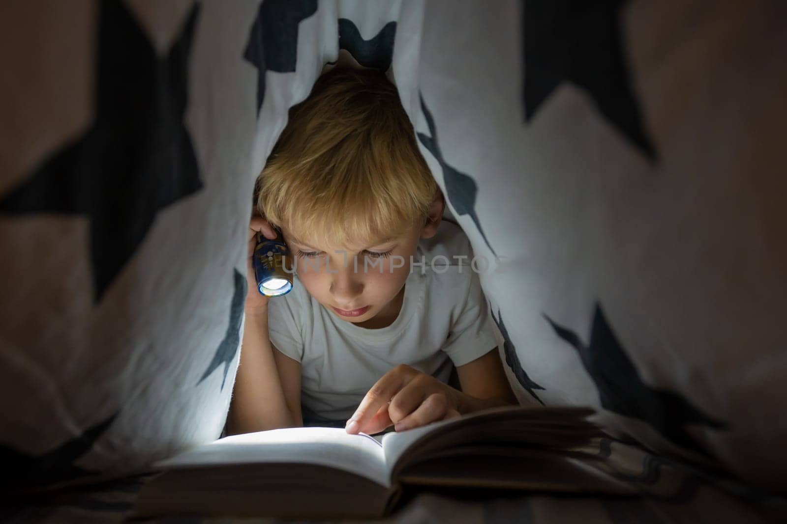 A little boy reads a book with a flashlight under the covers at night. by DovidPro