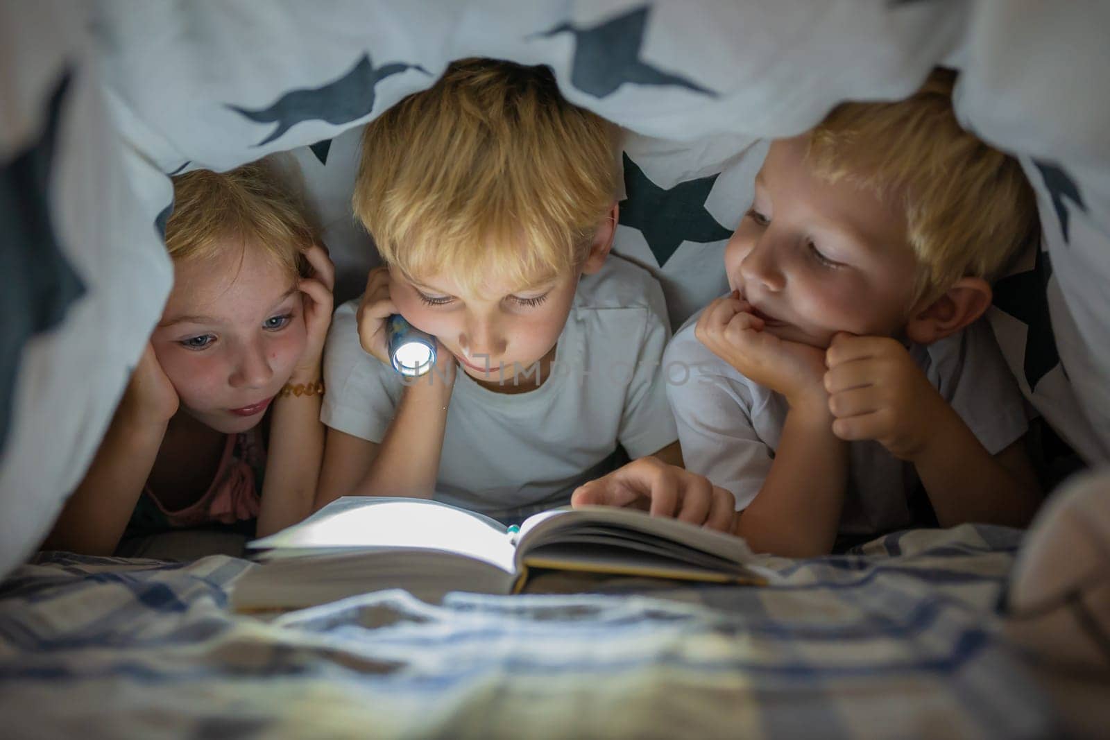 Three young children are reading a book with a flashlight under the covers at night