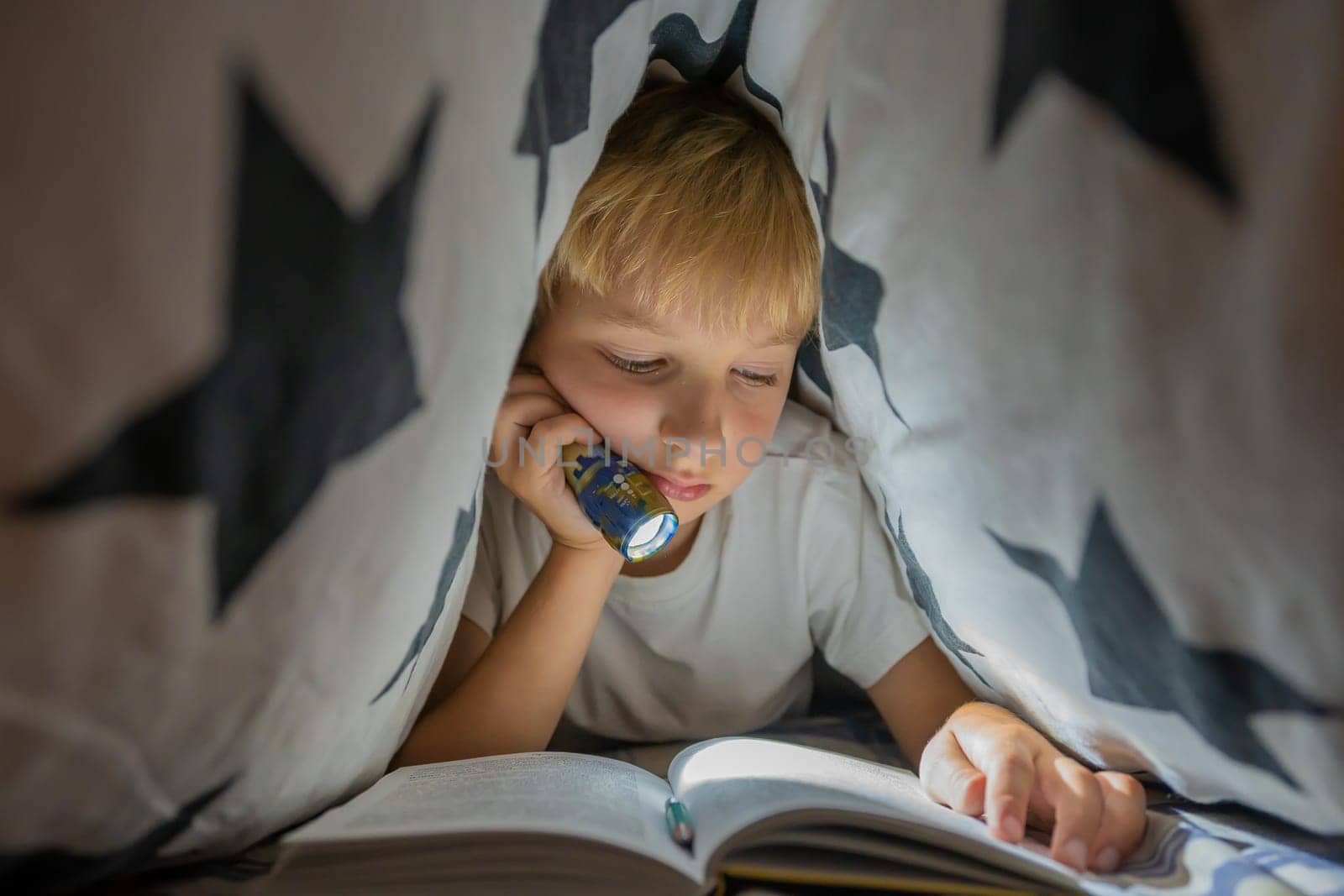 A little boy reads a book with a flashlight under the covers at night