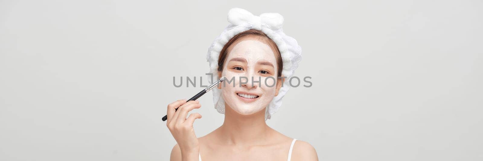 Beautiful young woman with clay facial mask, beauty treatment over white banner by makidotvn