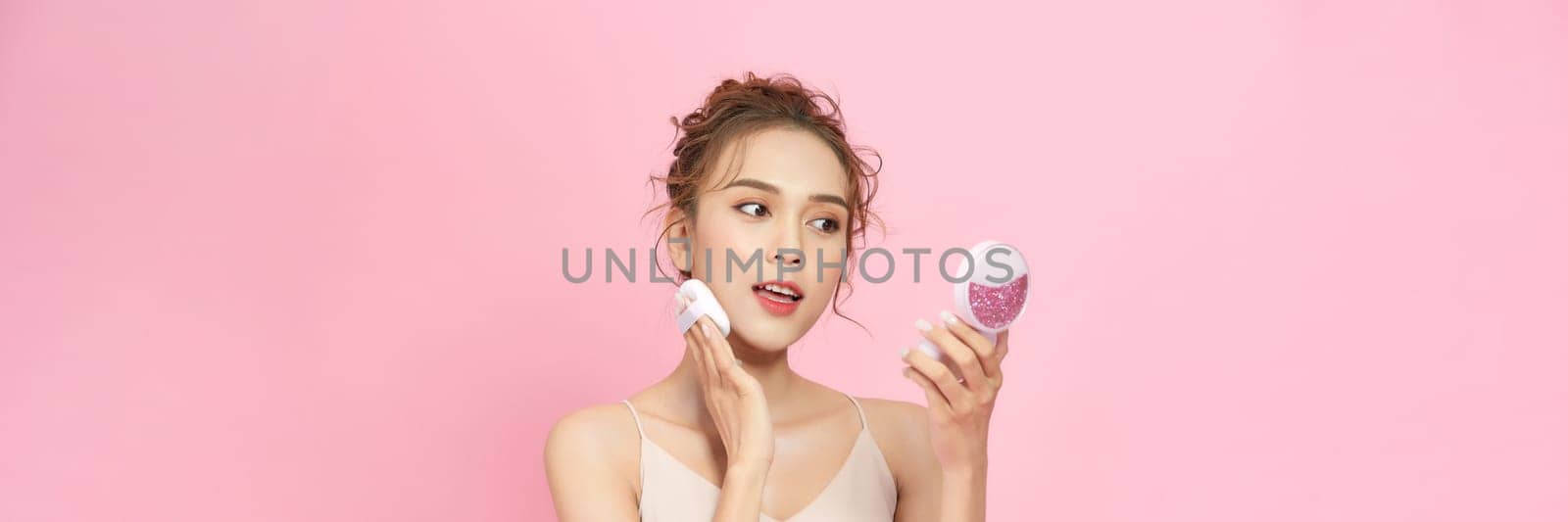 Portrait of attractive cheery girl applying blush tone foundation isolated over pink pastel background