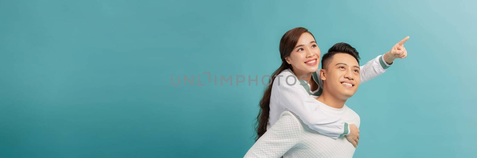 Happy Asian couple in love having fun piggyback ride and point at empty copyspace