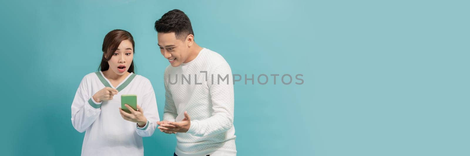 Amazed attractive young Asian couple looking at smartphone together isolated on blue background by makidotvn