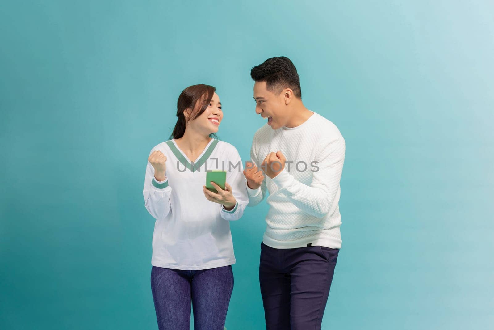 Excited young couple friends guy girl posing on blue background, holding using mobile phone