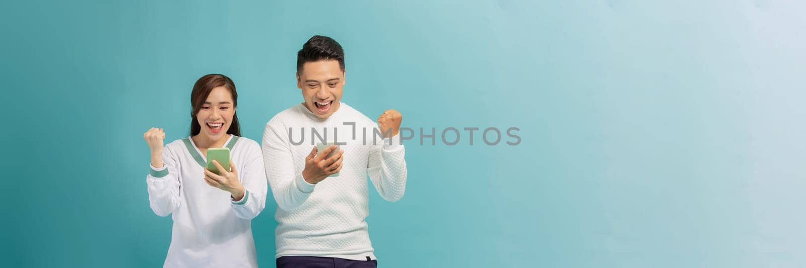 Cheerful attractive couple standing isolated over blue, using mobile phone, celebrating success by makidotvn