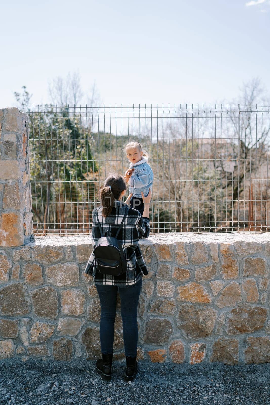 Mom holds a little girl standing on the stone base of a wire fence in the park. Back view. High quality photo