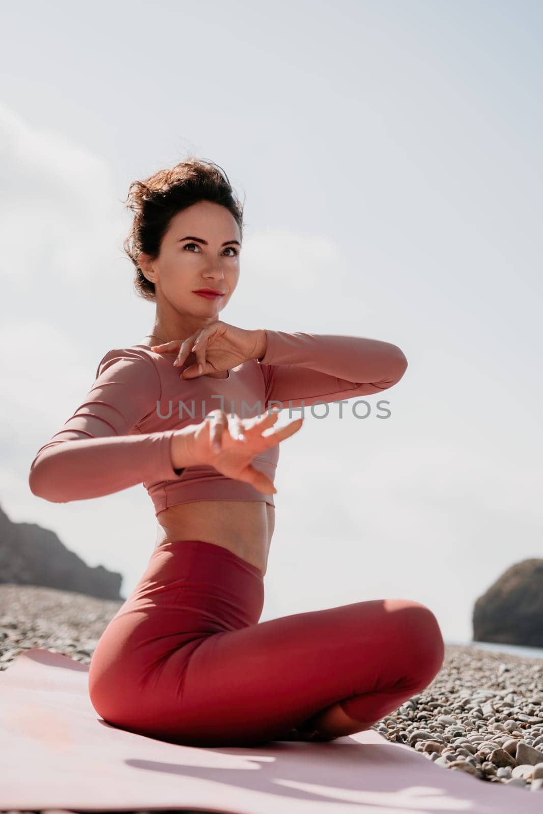 Middle aged well looking woman with black hair, fitness instructor in leggings and tops doing stretching and pilates on yoga mat near the sea. Female fitness yoga routine concept. Healthy lifestyle by panophotograph