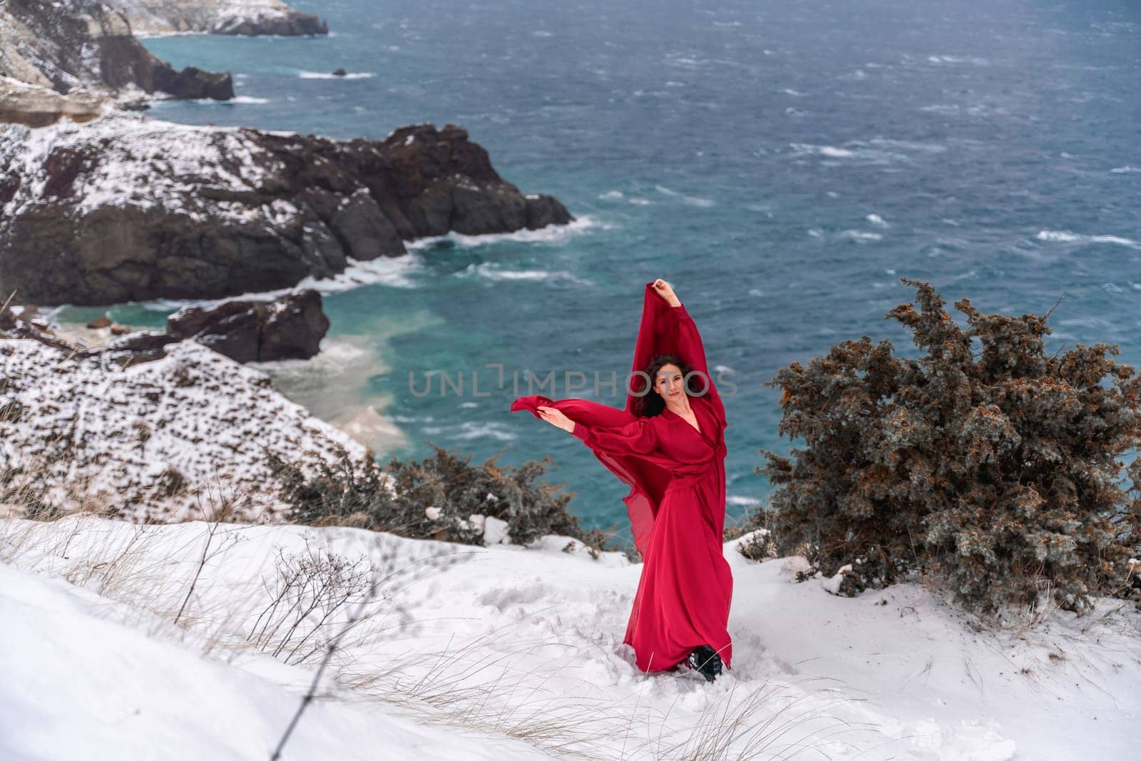 Woman red dress snow sea. Happy woman in a red dress in the snowy mountains by the emerald sea. The wind blows her clothes, posing against sea and snow background