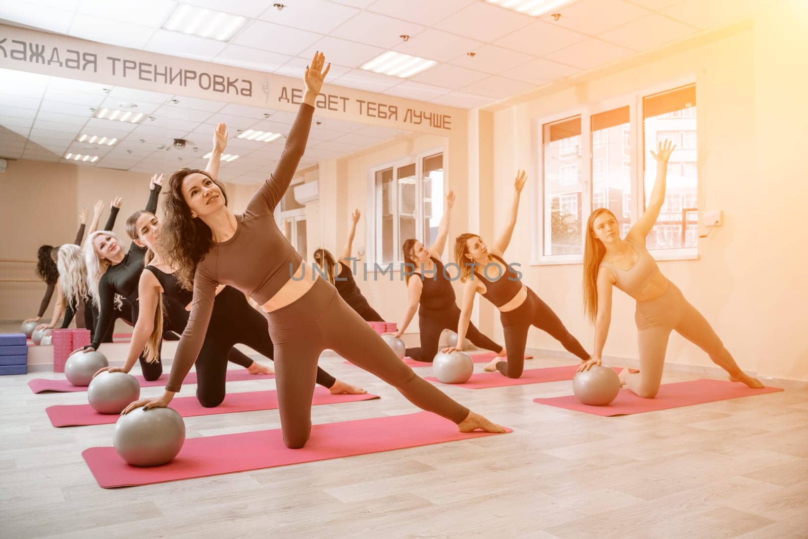 A group of six athletic women doing pilates or yoga on pink mats in front of a window in a beige loft studio interior. Teamwork, good mood and healthy lifestyle concept. by Matiunina