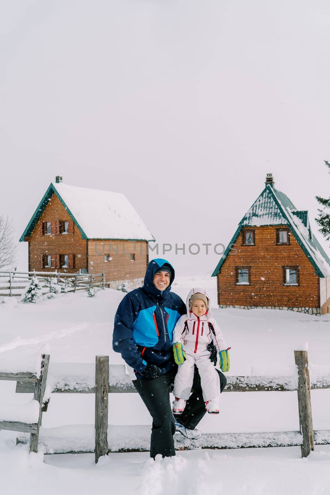 Smiling dad with a little girl on his lap sits on a fence in a village under snowfall. High quality photo