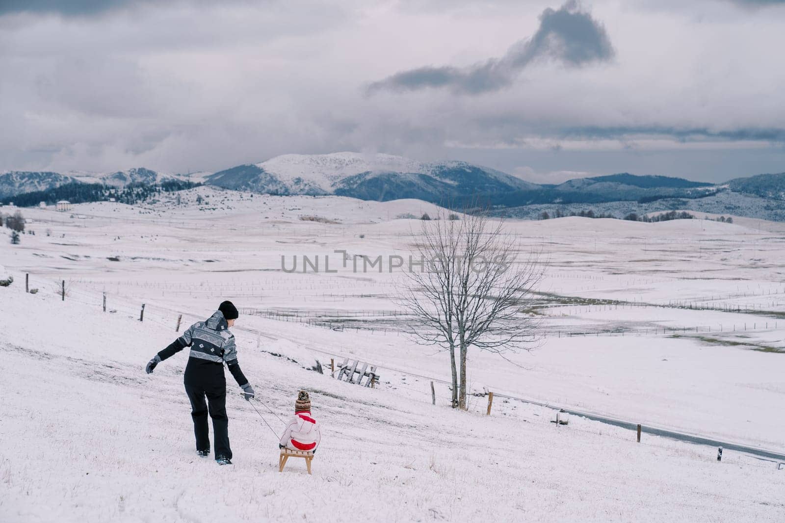 Mother carries a small child on a wooden sleigh along a snowy hill in a mountain valley. Back view. High quality photo