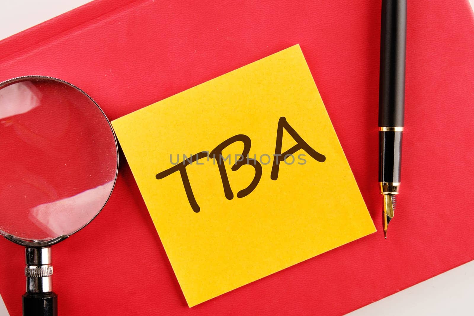 TBA the inscription on the sticker lying on a red notebook next to a magnifying glass and a fountain pen