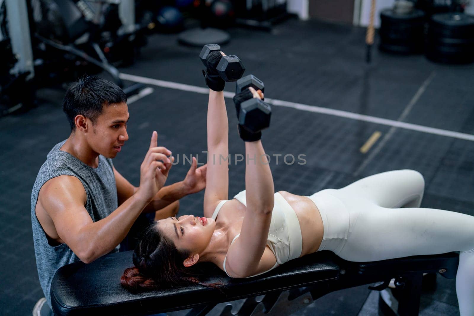 Asian coach help and support Asian sport woman lift dumbells up with lie position on long chair in fitness gym.