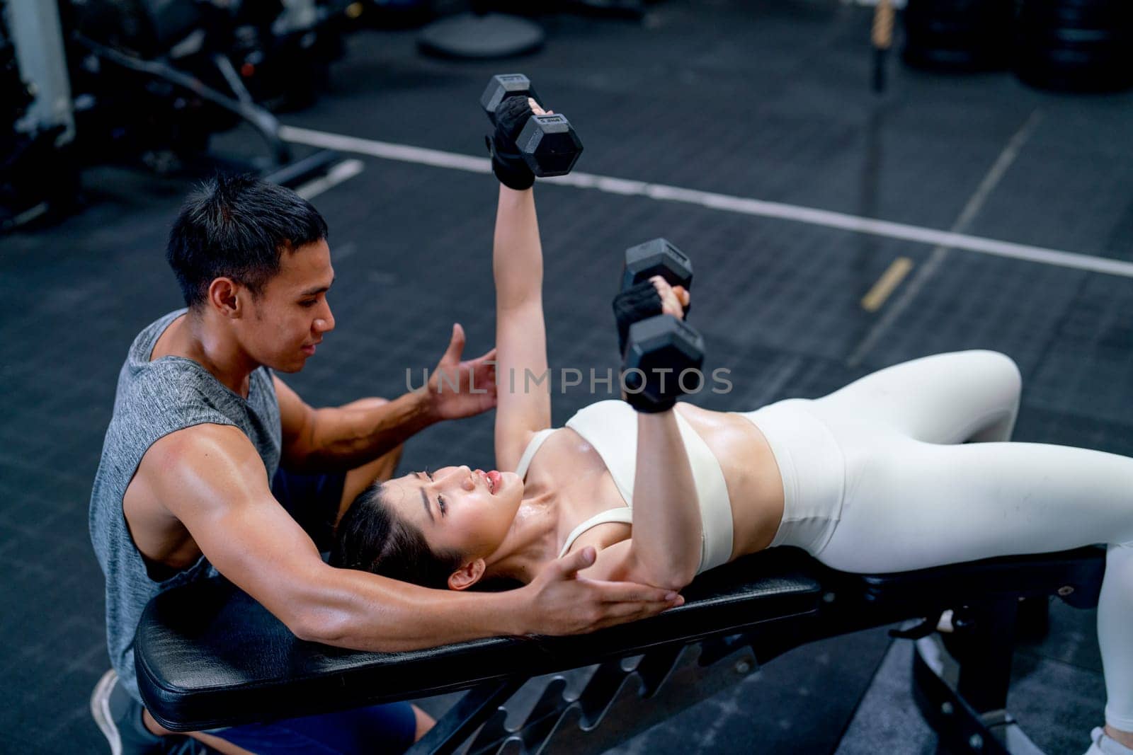 Asian coach help and support Asian sport woman lift dumbells up with lie position on long chair in fitness gym.