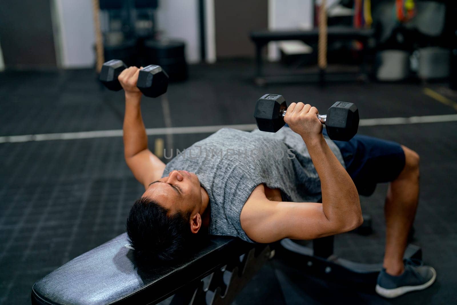 Top view of Asian sport man lift dumbells up to over head during exercise and lie on long chair in fitness gym.