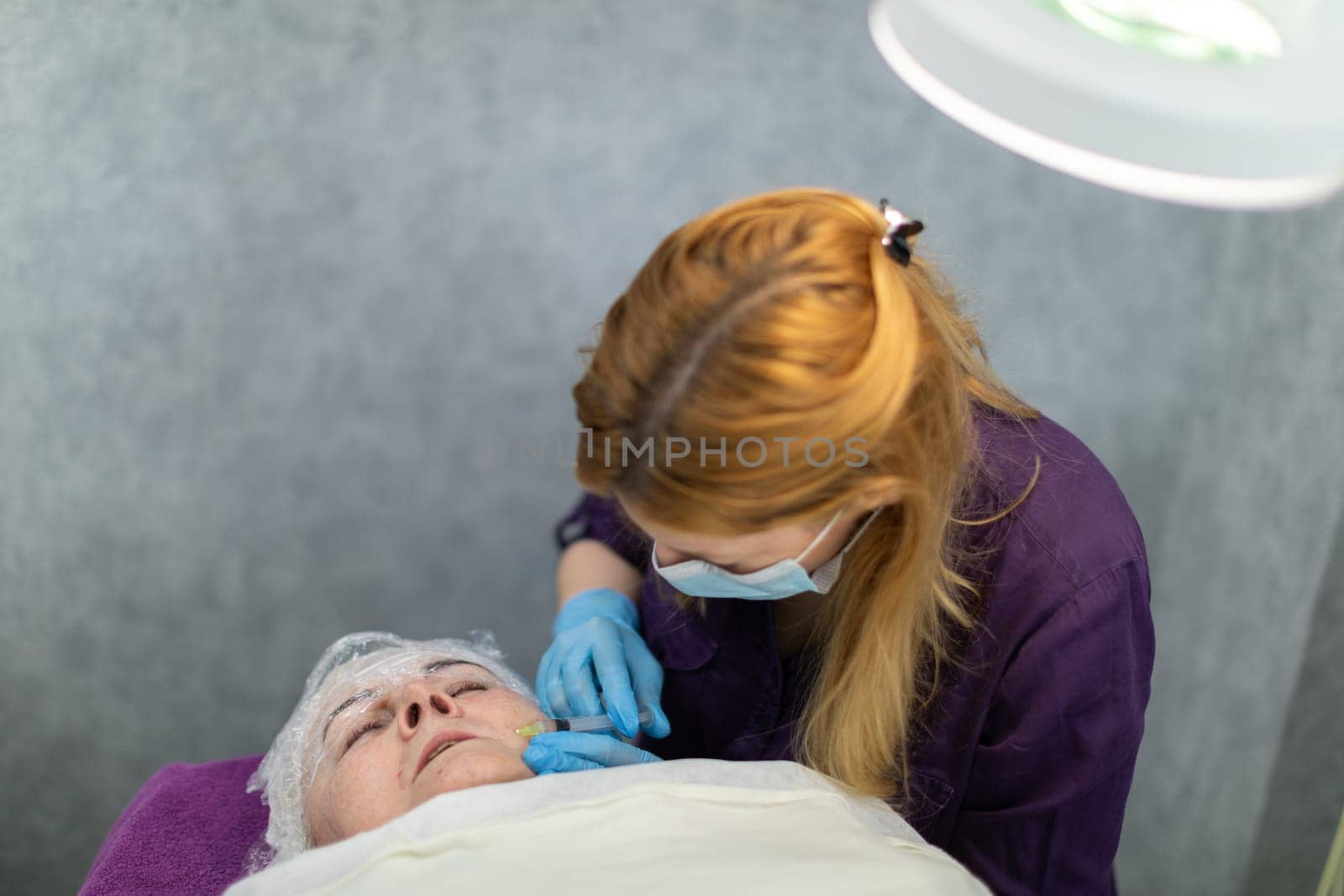 The patient is lying on a bed in an aesthetic clinic, with a lamp lighting over her. The woman has a grimace of pain on her face as she has needle mesotherapy performed.