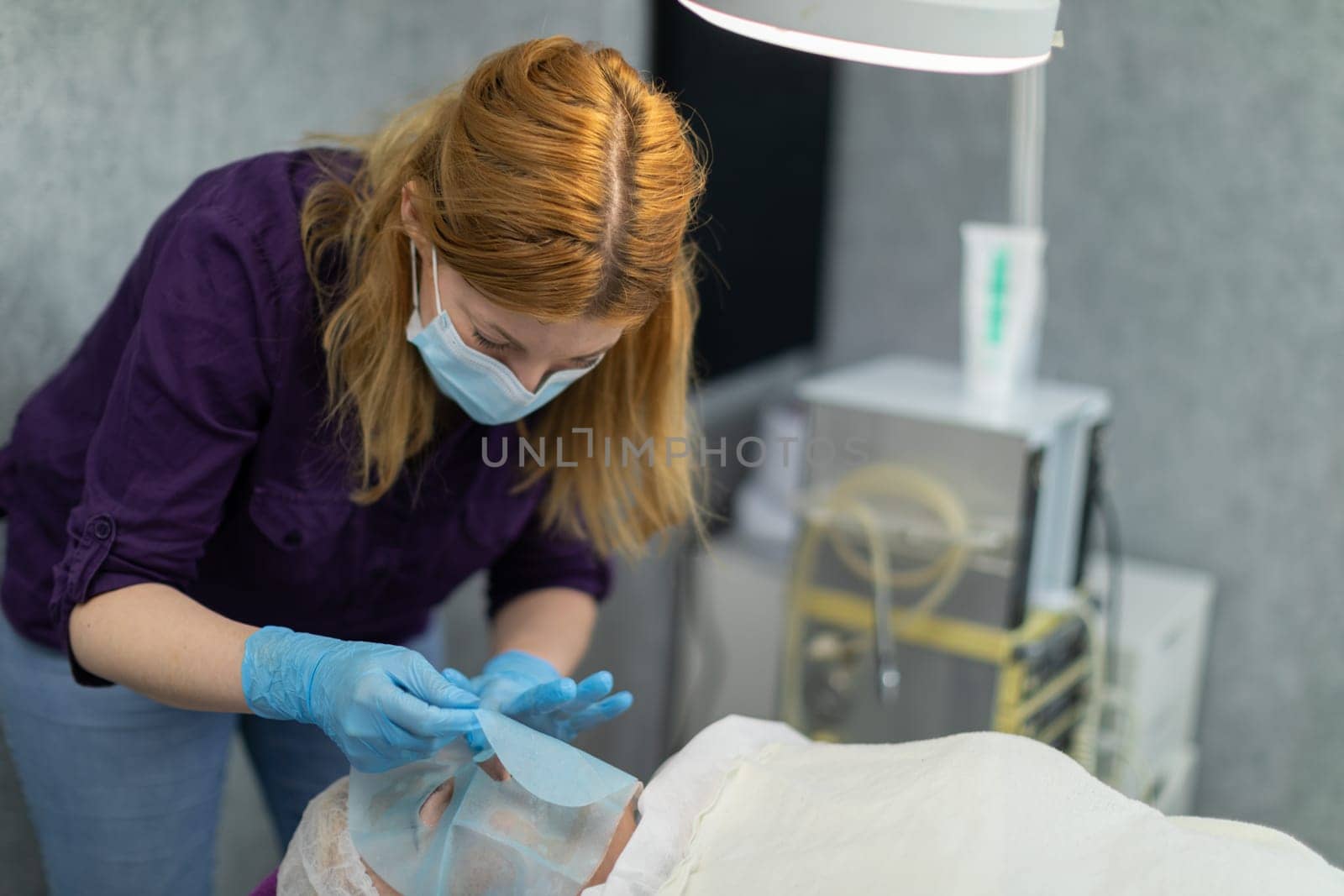 A beautician performs a facial moisturizing service for a lying woman. The client of the beauty salon is lying on a bed, covered with a white sheet.