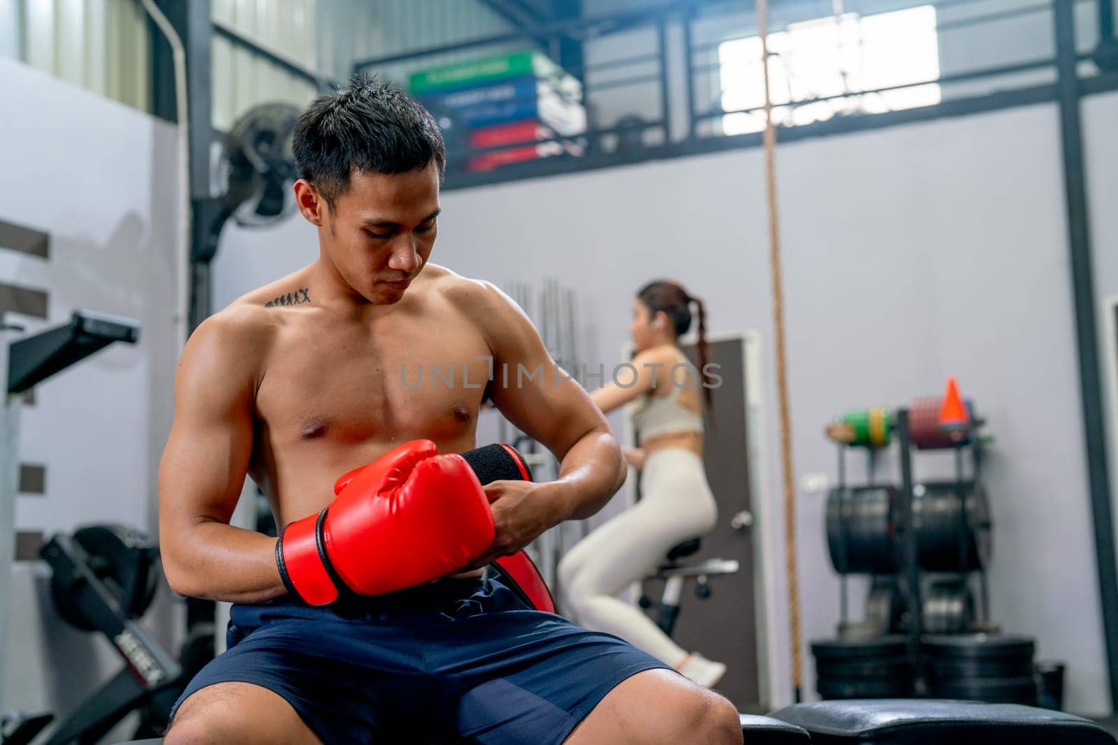 Asian sport man sit on chair and try to wear boxing glove in front of the woman exercise in the back of fitness gym.