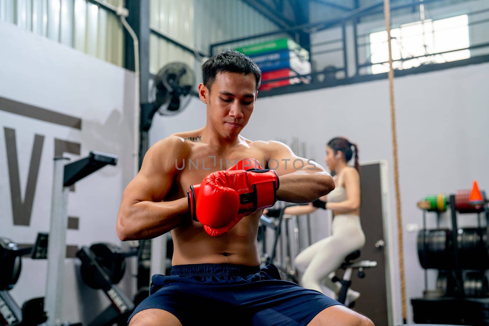 Asian sport man sit on chair and try to wear boxing glove in front of the woman exercise in the back of fitness gym.