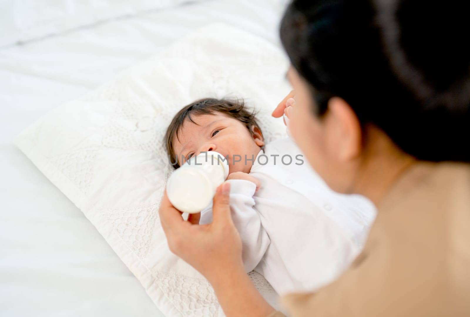 Back of Young Asian mother give milk in bottle to newborn baby lie on bed and take care with happiness in bedroom of their house.